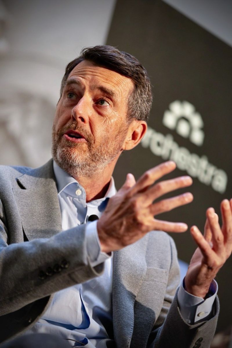 A speaker gesturing in front of an Orchestra backdrop